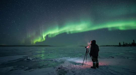 Une personne observe des aurores boréales depuis un lac gelé au Canada, appareil photo sur trépied et lampe frontale allumée.