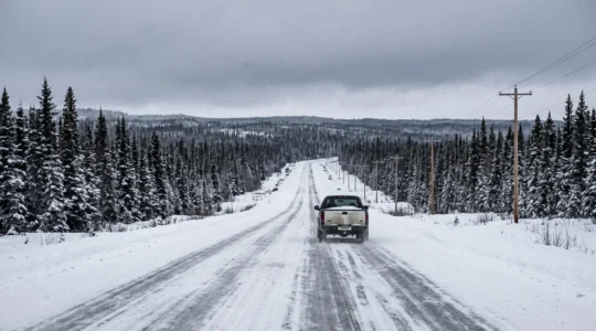 Une route enneigée traverse une immense forêt canadienne, avec un seul véhicule au loin sous un ciel gris.